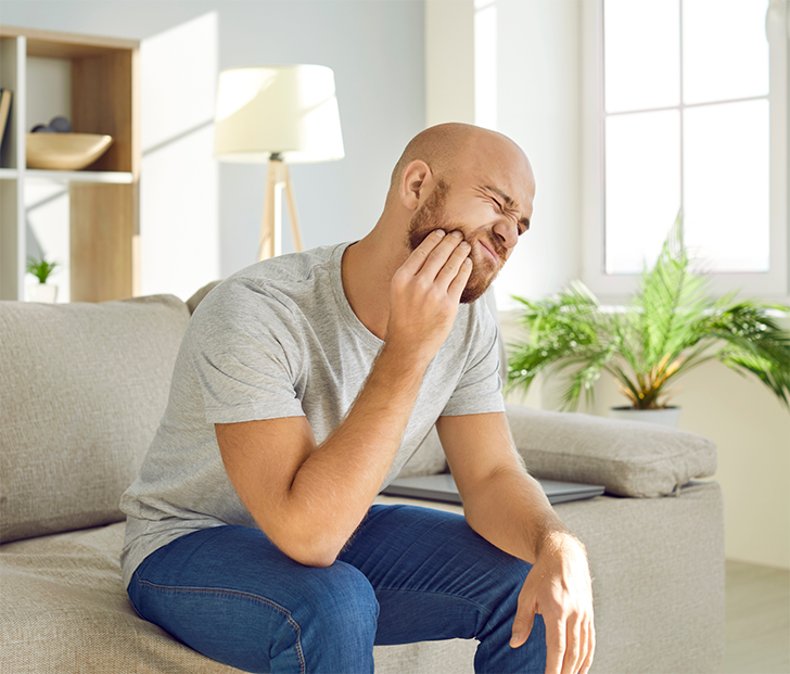 Bearded man sitting on couch and rubbing jaw in pain
